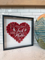 Red paper roses in a heart with the couple's names in white script, in a black shadow box frame