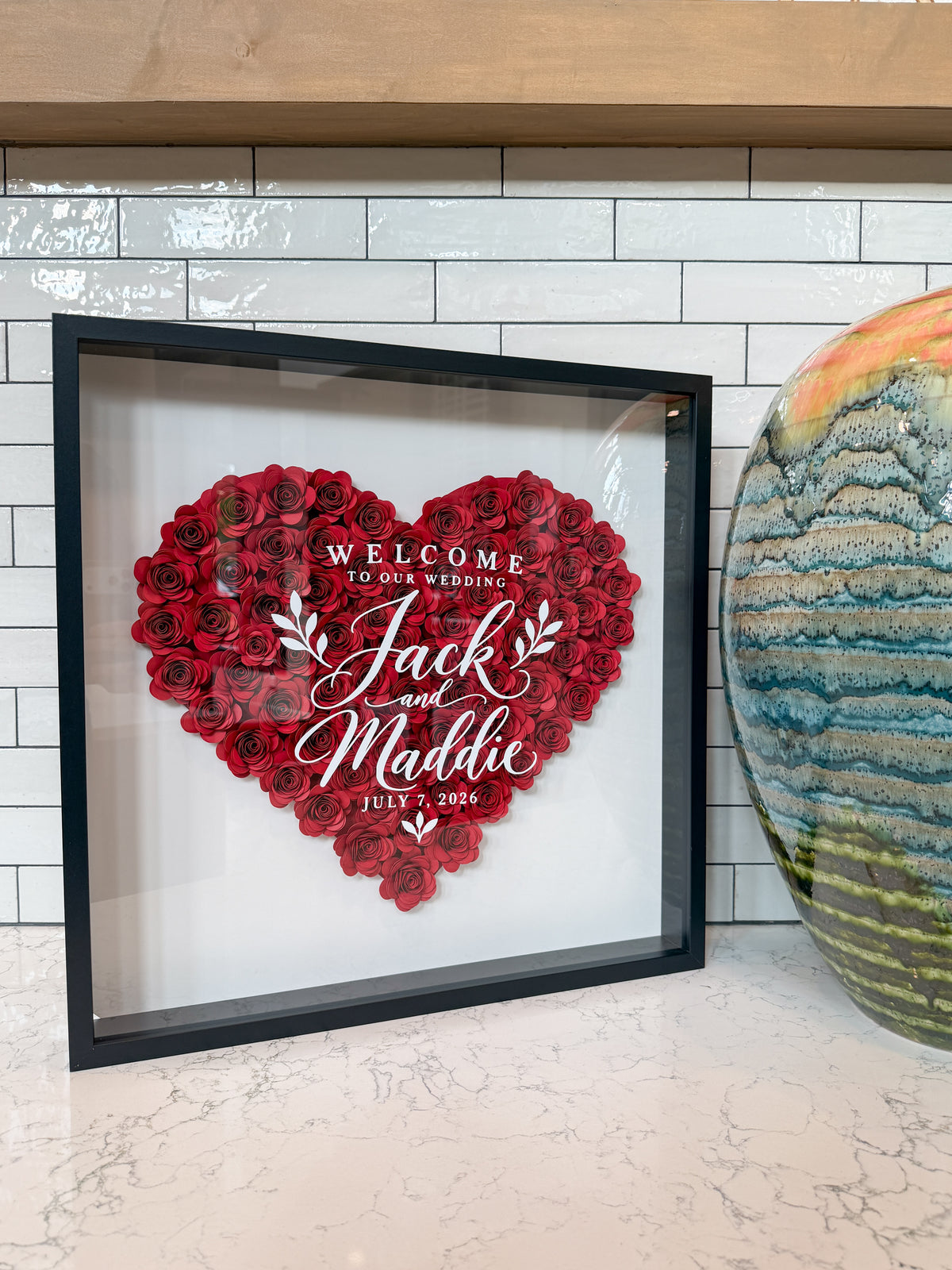 Red paper roses in a heart with the couple's names in white script, in a black shadow box frame