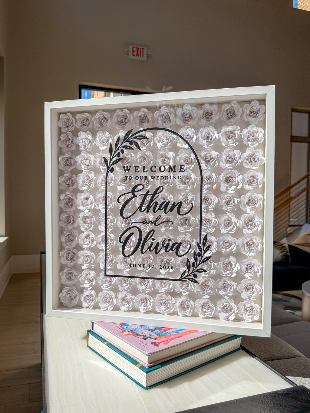 White frame filled edge to edge with white paper roses, names lettered in black on the glass